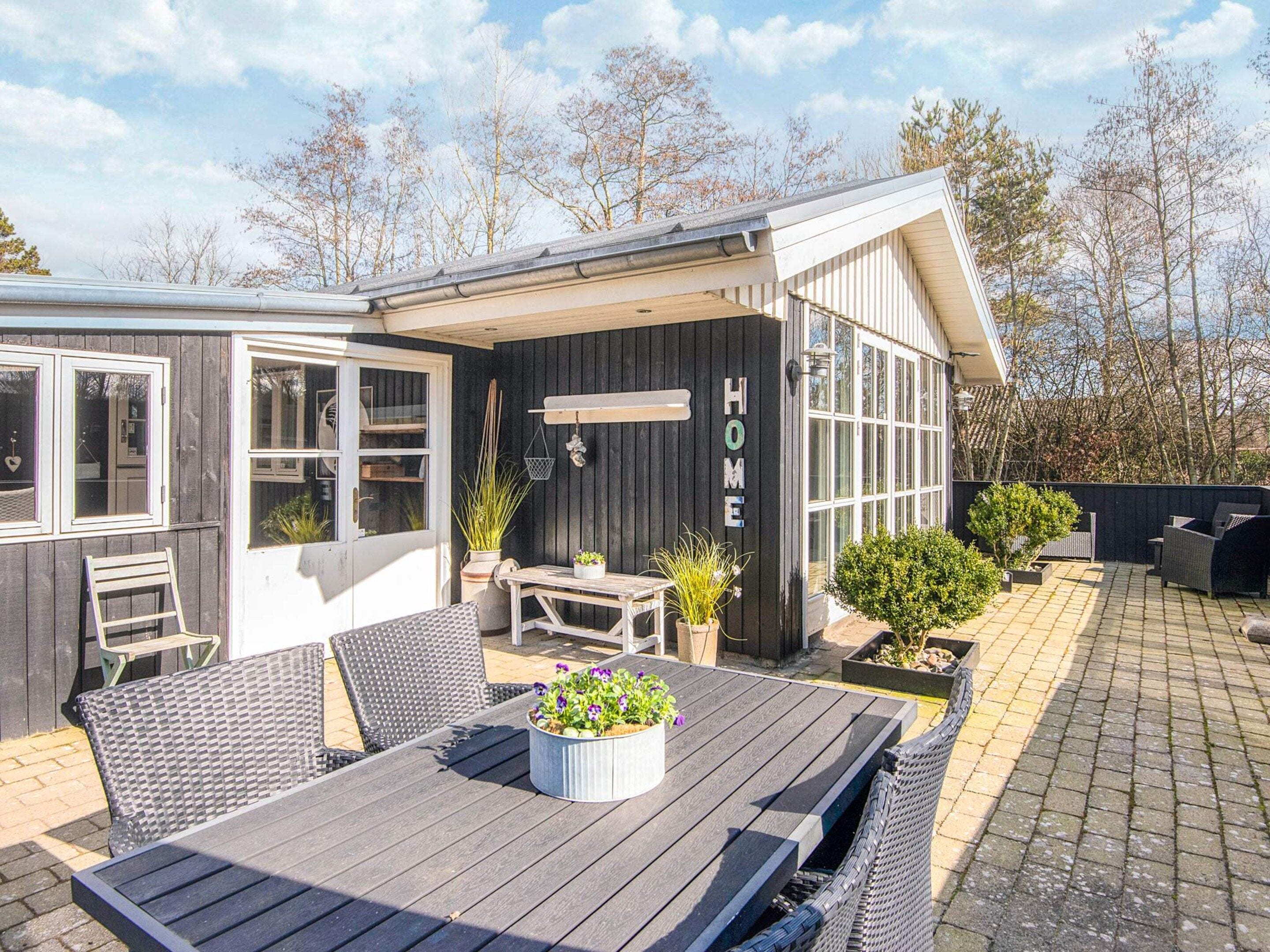 Une terrasse avec une table et des chaises, un petit jardin et un bâtiment sur lequel on peut lire le mot « MAISON ».