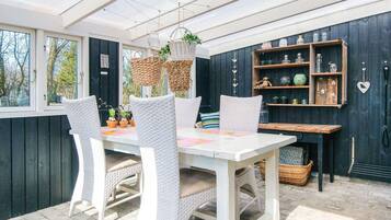 A covered outdoor dining area with wicker chairs and a table, surrounded by potted plants and a wooden shelf with decorative items.