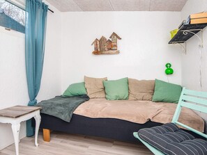 A cozy bedroom with a bed, a small table, a chair, and a shelf with books and a green lamp.