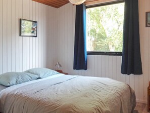 A bedroom with a bed, a nightstand, a lamp, a window with curtains, and a picture on the wall.
