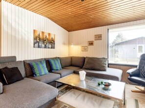 A modern living room with a wooden ceiling, a grey sectional sofa with colorful cushions, a glass coffee table, and a large window with blinds. - 6 Person Holiday Home in Vaeggerlose (Vaeggerlose)