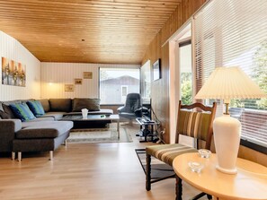 A modern living room with a wooden ceiling, a sofa set, a dining table with a lamp, and a view through large windows. - 6 Person Holiday Home in Vaeggerlose (Vaeggerlose)