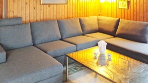 A room with a gray sectional sofa, a glass coffee table with a vase, and wooden panel walls.