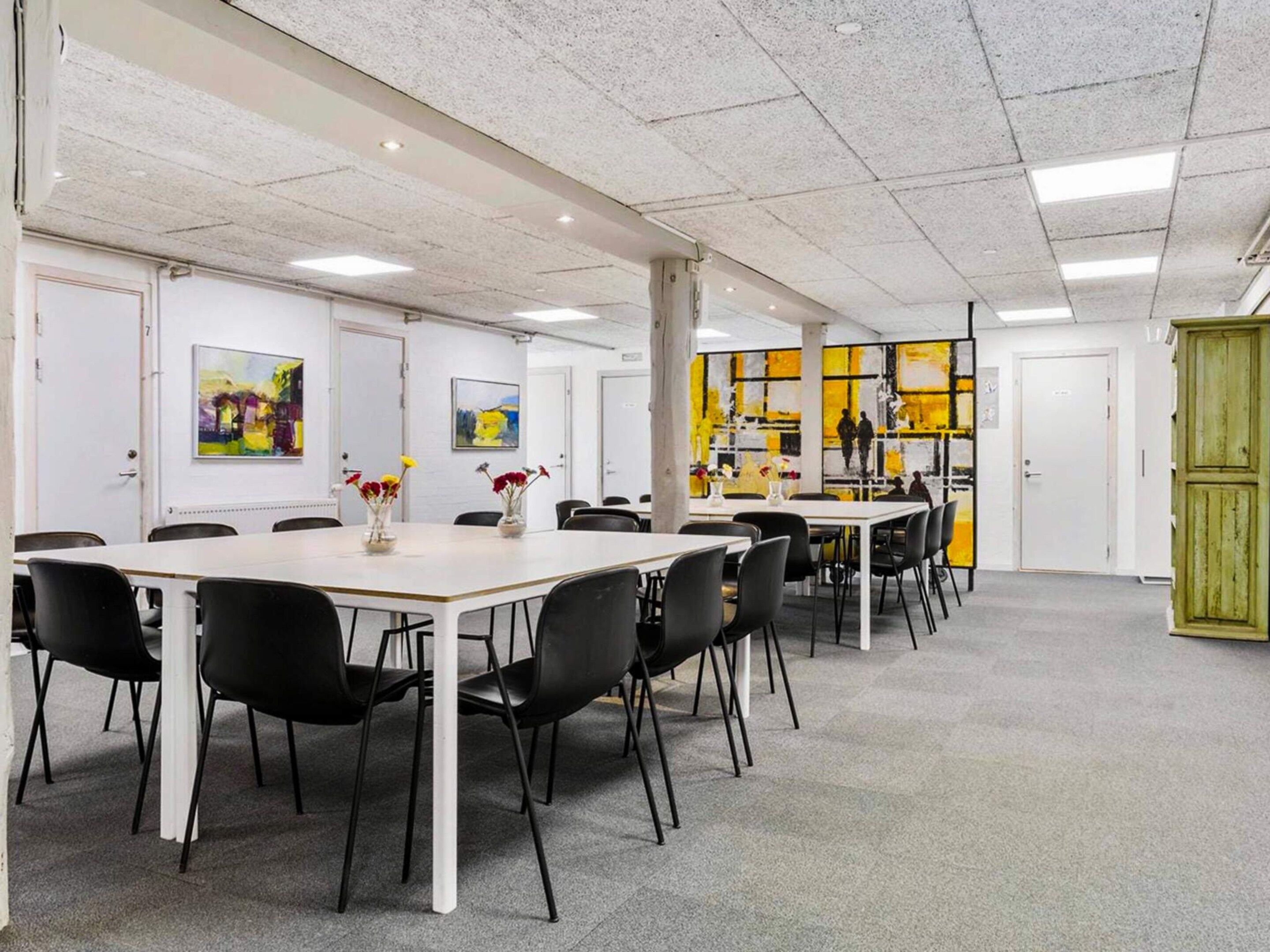 Una sala conferenze con tavoli bianchi e sedie nere, una lavagna e un quadro astratto giallo e nero appeso al muro.