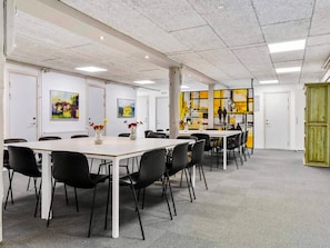 A spacious conference room with white tables and black chairs, featuring artwork on the walls and a green cabinet in the corner.