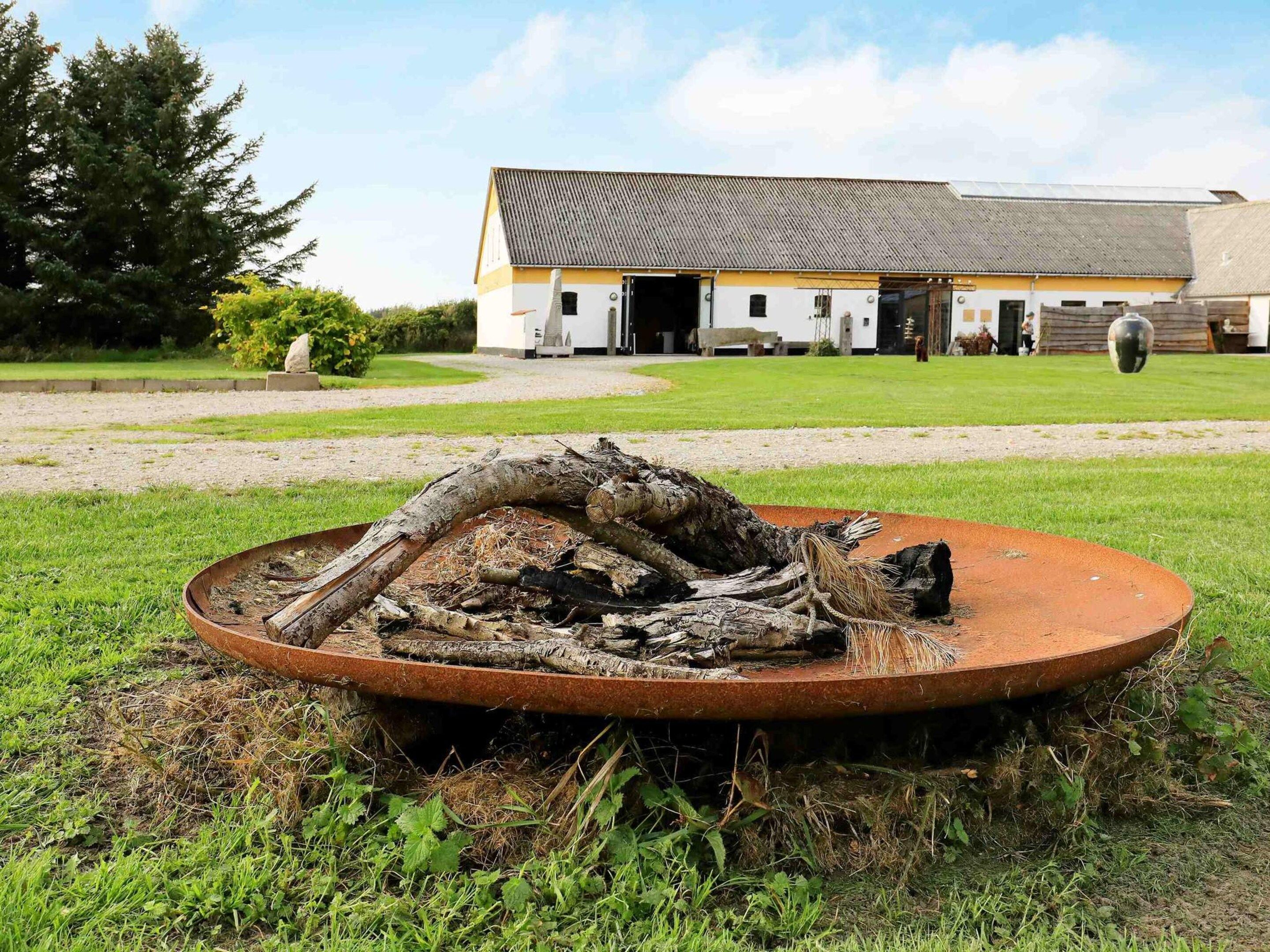 A rustic outdoor area with a large metal fire pit surrounded by logs and branches. In the background, there is a building with a sloped roof and a paved driveway.