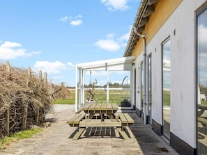 A covered outdoor seating area with a table and benches, adjacent to a building with large windows.