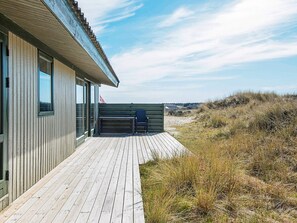 A wooden deck leading to a building with large windows, a bench, and a view of the beach and dunes.