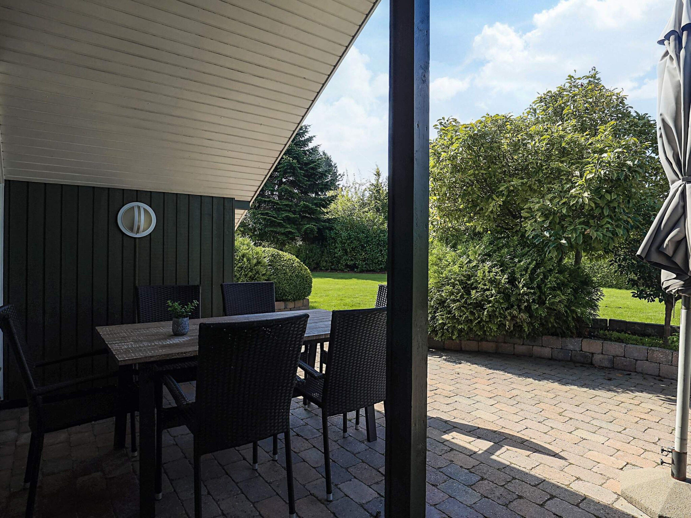 An outdoor patio with a table and chairs, a covered area, and a view of a well-maintained garden and trees.