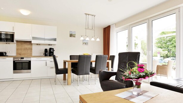 A modern kitchen with white cabinets, a dining area with a wooden table and black chairs, and a large window with a view of the outdoors. - Holiday Home in GroÃŸenbrode (Grossenbrode)