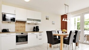 A modern kitchen with white cabinetry, a built-in oven, a microwave, and a coffee maker. There is a dining table with four chairs and a vase with flowers. - Holiday Home in GroÃŸenbrode (Grossenbrode)