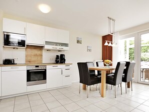 A modern kitchen with white cabinetry, a built-in oven, a microwave, and a coffee maker. There is a dining table with four chairs and a vase with flowers. - Holiday Home in GroÃŸenbrode (Grossenbrode)