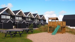 Une rangée de maisons de plage noires avec des bordures blanches, une structure de jeu en bois avec un toboggan et des tables de pique-nique.