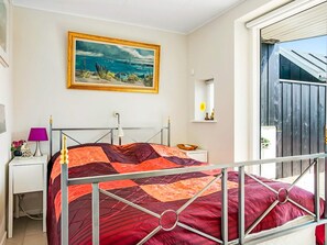 A bedroom with a metal bed frame, a painting of a coastal scene, a bedside table with a lamp, and a window with a view of the outside.