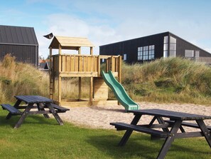 A playground with a slide and a wooden climbing structure, picnic tables, and a building in the background.