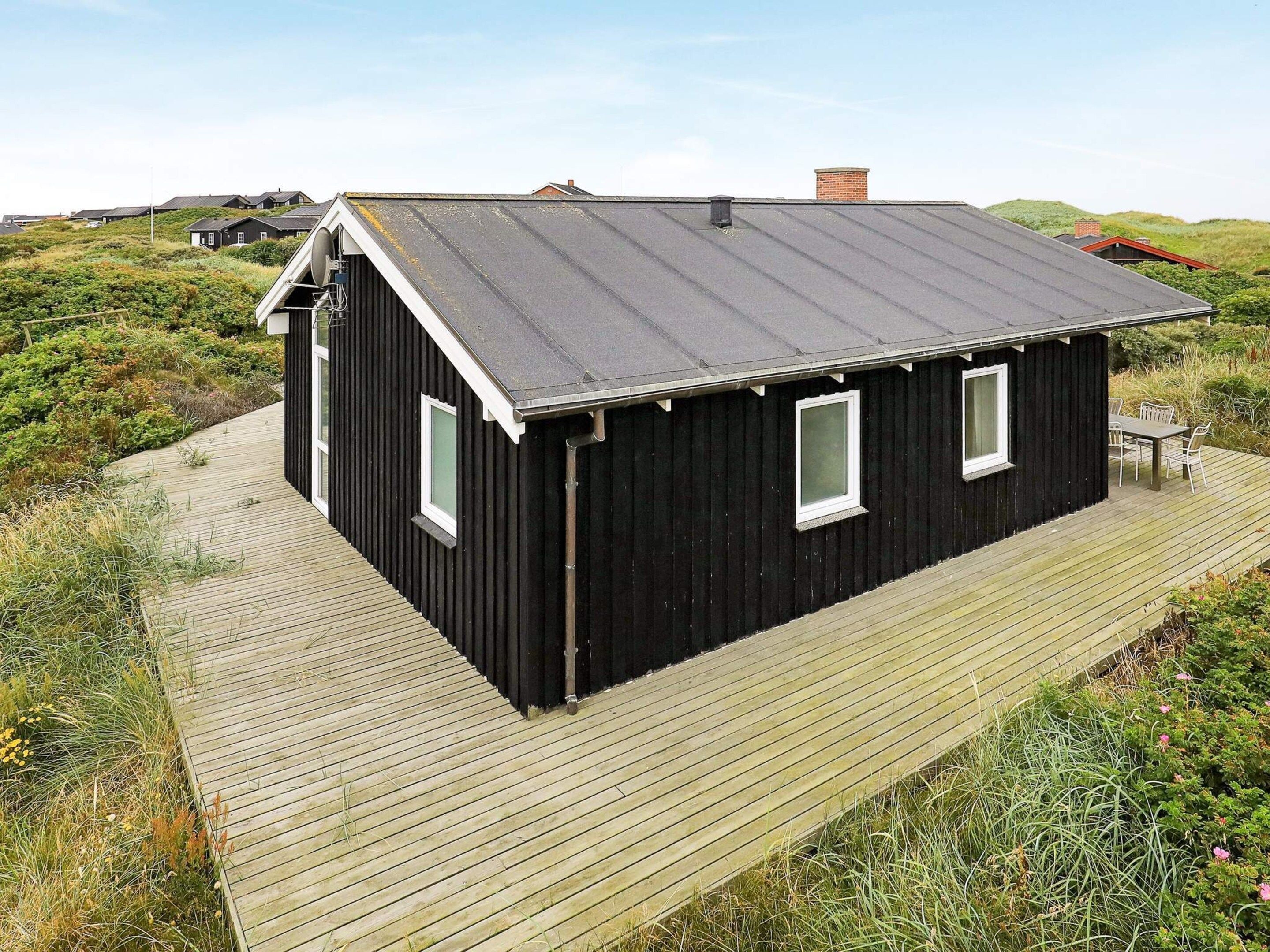 A black wooden house with a sloped roof, a wooden deck, and a small outdoor seating area.