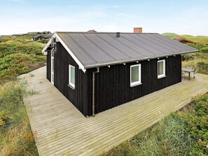 Une maison en bois noire, avec un toit en pente, une terrasse en bois et un petit espace extérieur aménagé pour s’asseoir.
