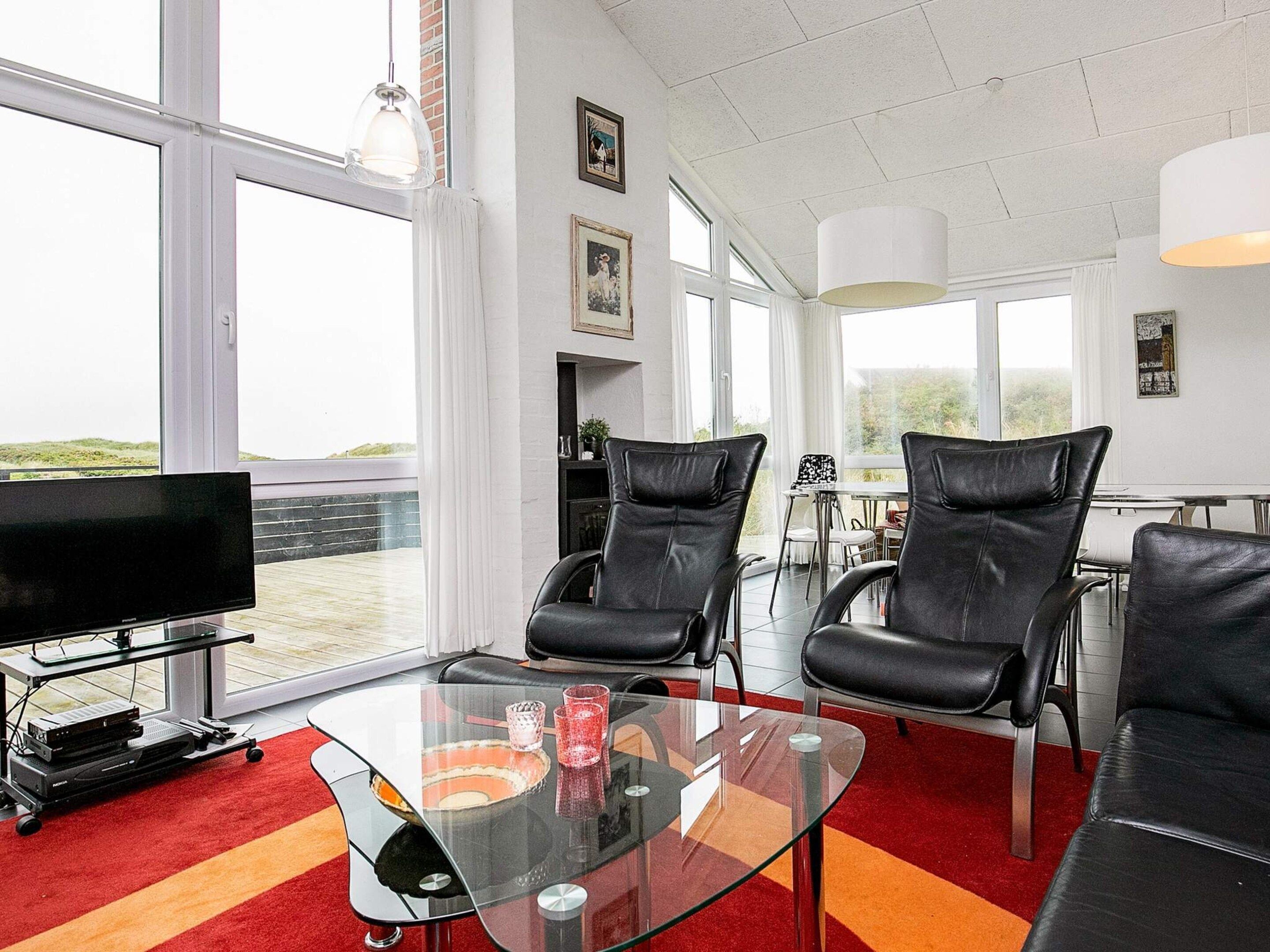 A modern living room with a large TV, leather chairs, a glass coffee table, and large windows offering a view.