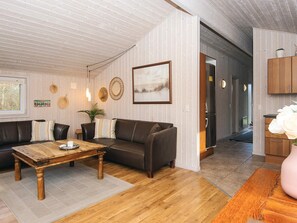 A modern living room with a wooden coffee table, leather sofa, and a dining table. There is a kitchen area with wooden cabinets and a refrigerator.