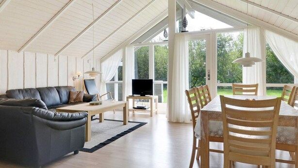 Amplia sala de estar con un sofá de cuero negro, una mesa de comedor de madera y sillas. Un televisor se encuentra sobre un mueble de madera, y la habitación cuenta con grandes ventanas que dejan entrar luz natural.