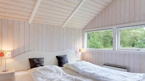 Un dormitorio con techo de madera, una cama grande con ropa de cama blanca, dos almohadas, una mesita de noche con una lámpara y una ventana con vistas a árboles.