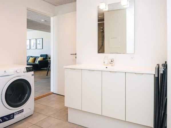 A laundry room with a washing machine, a sink with a mirror, and a door leading to a living area with a couch and a coffee table.