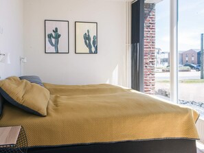A bed with a yellow blanket, two framed cactus pictures on the wall, and a view of a brick building through a window.