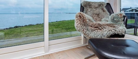 Una habitación con un ventanal grande que da a una masa de agua, un sillón cubierto de piel y un banco de cuero negro.