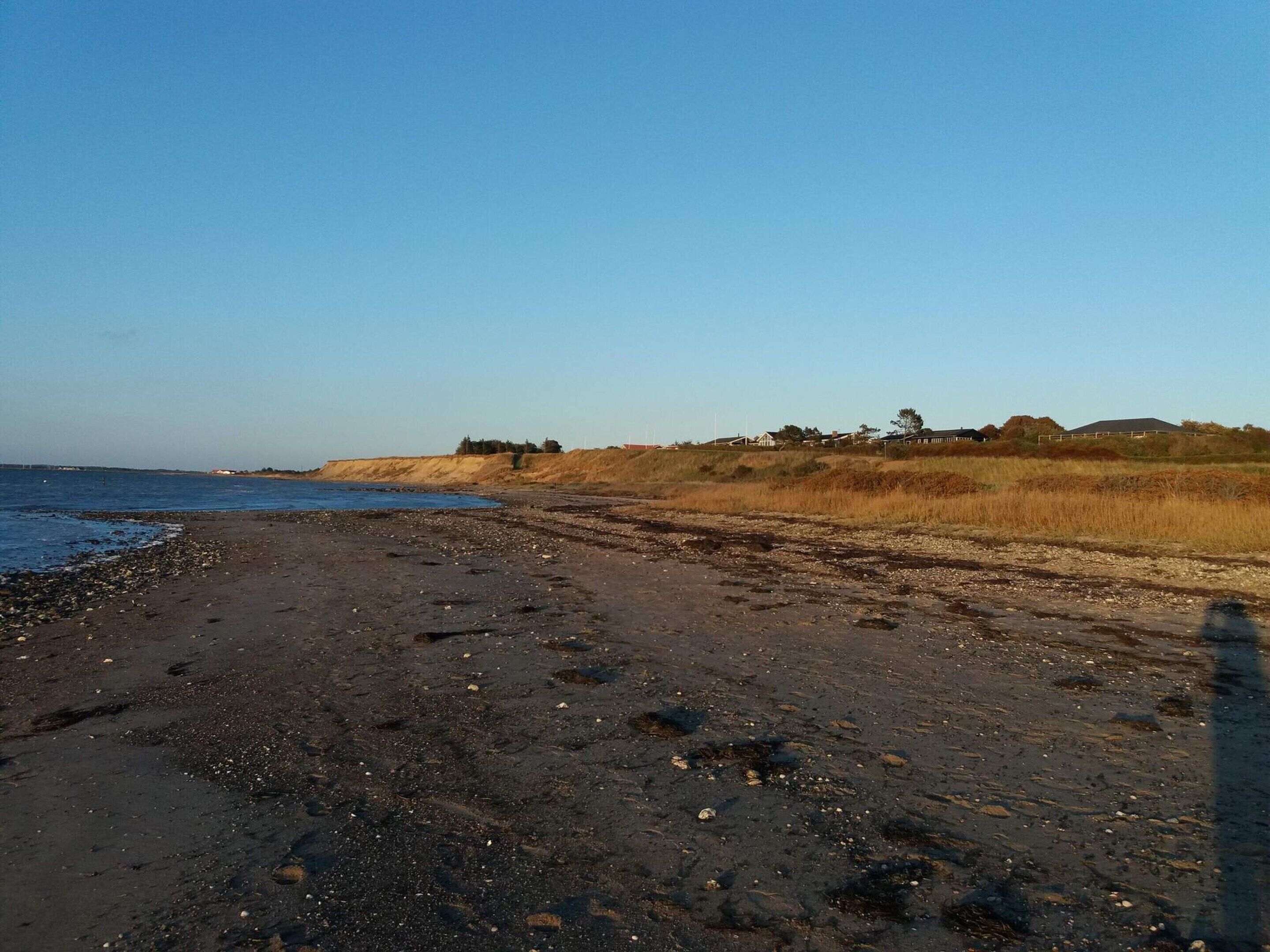 A beach with a clear sky, a sandy shore, and a distant view of a hill with sparse vegetation.