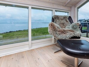A room with a large window overlooking a body of water, a chair with a fur throw, and a wooden floor. - 5 Star Holiday Home in Vinderup (Vinderup)