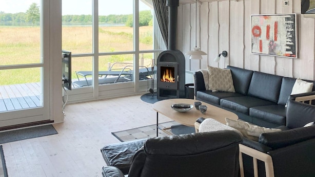 A cozy living room with a fireplace, a black leather sofa, a coffee table, and a view of a grassy field through large windows.