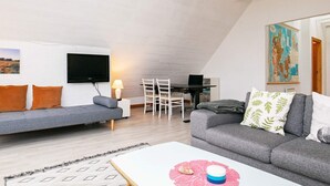 A modern loft-style living room with a high ceiling, a flat-screen TV mounted on the wall, a grey sofa with orange cushions, a grey couch, a small dining table with chairs, and a map on the wall. - 9 Person Holiday Home in Thisted-by Traum (Thisted)