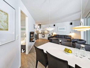 Eine moderne Küche mit weißen Schränken, ein Essbereich mit einem weißen Tisch und ein Wohnbereich mit einem dunkelgrauen Sofa.
