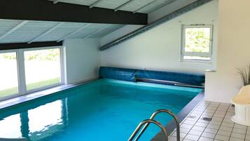 An indoor swimming pool with clear water, a metal ladder, and a window with a view of greenery.