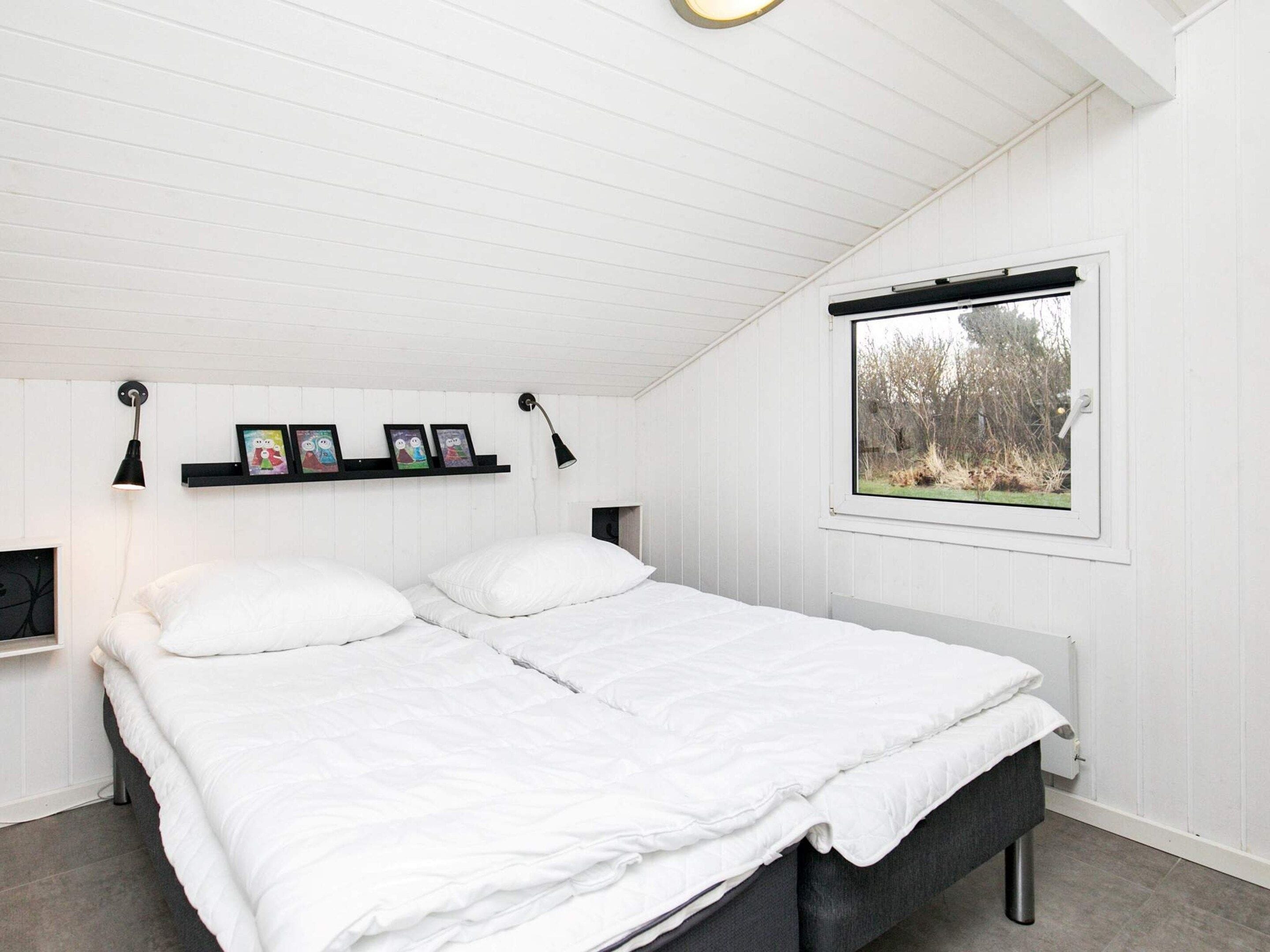 A bedroom with a bed, two bedside lamps, a shelf with framed pictures, and a window with a view of trees.