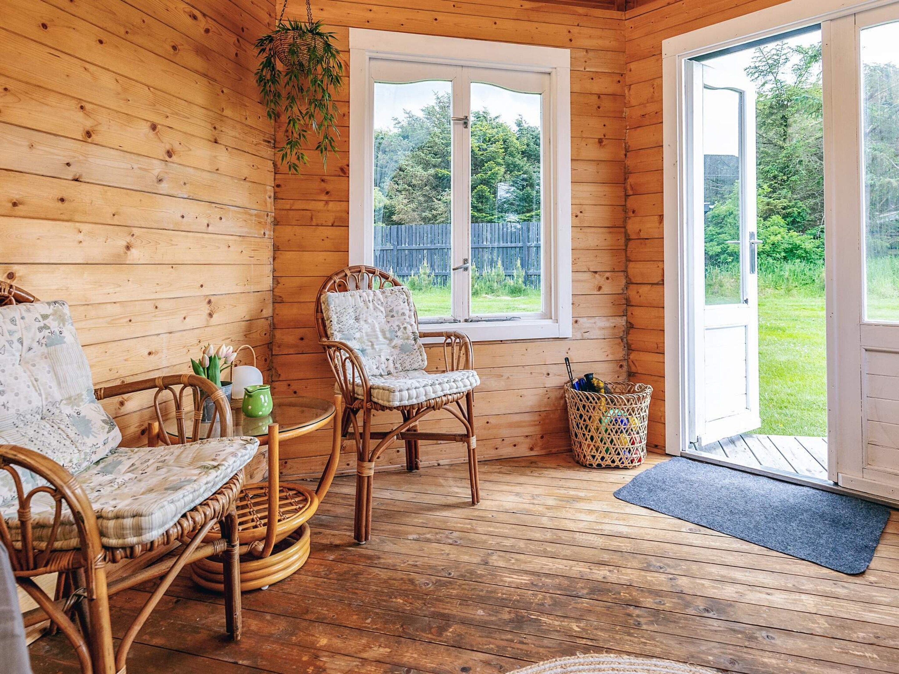 Interior de una cabaña de madera con muebles de mimbre, una ventana con vistas a un paisaje verde y una puerta que da al exterior.