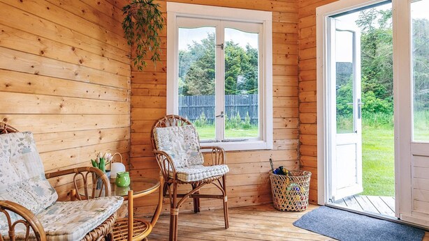 A wooden cabin interior with wicker furniture, a window with a view of a green landscape, and a door leading outside.