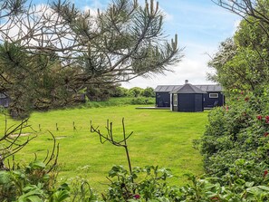 A small, dark-colored building with a sloped roof, situated in a grassy area with trees and shrubs around. - Lovely Holiday Home in LÃ¸nstrup near Sea (Hjørring)