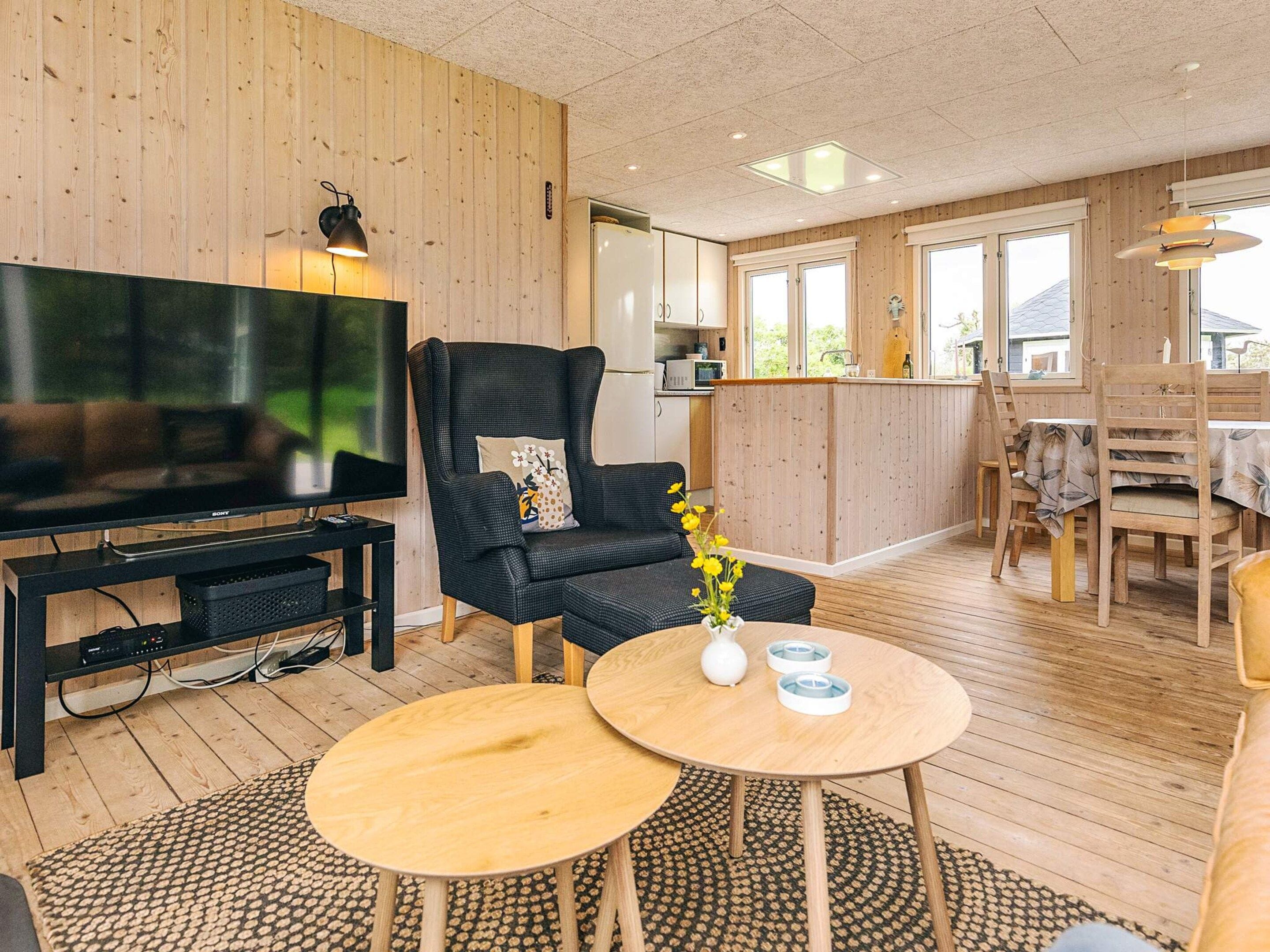 En moderne stue med et fladskærms-tv, en sort læderlænestol, et træspisebord og et køkken i baggrunden.