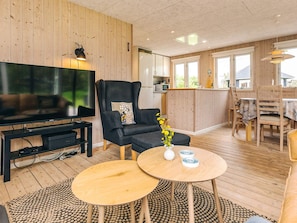A modern living room with a flat-screen TV, a black leather armchair, a wooden dining table, and a kitchen area in the background.