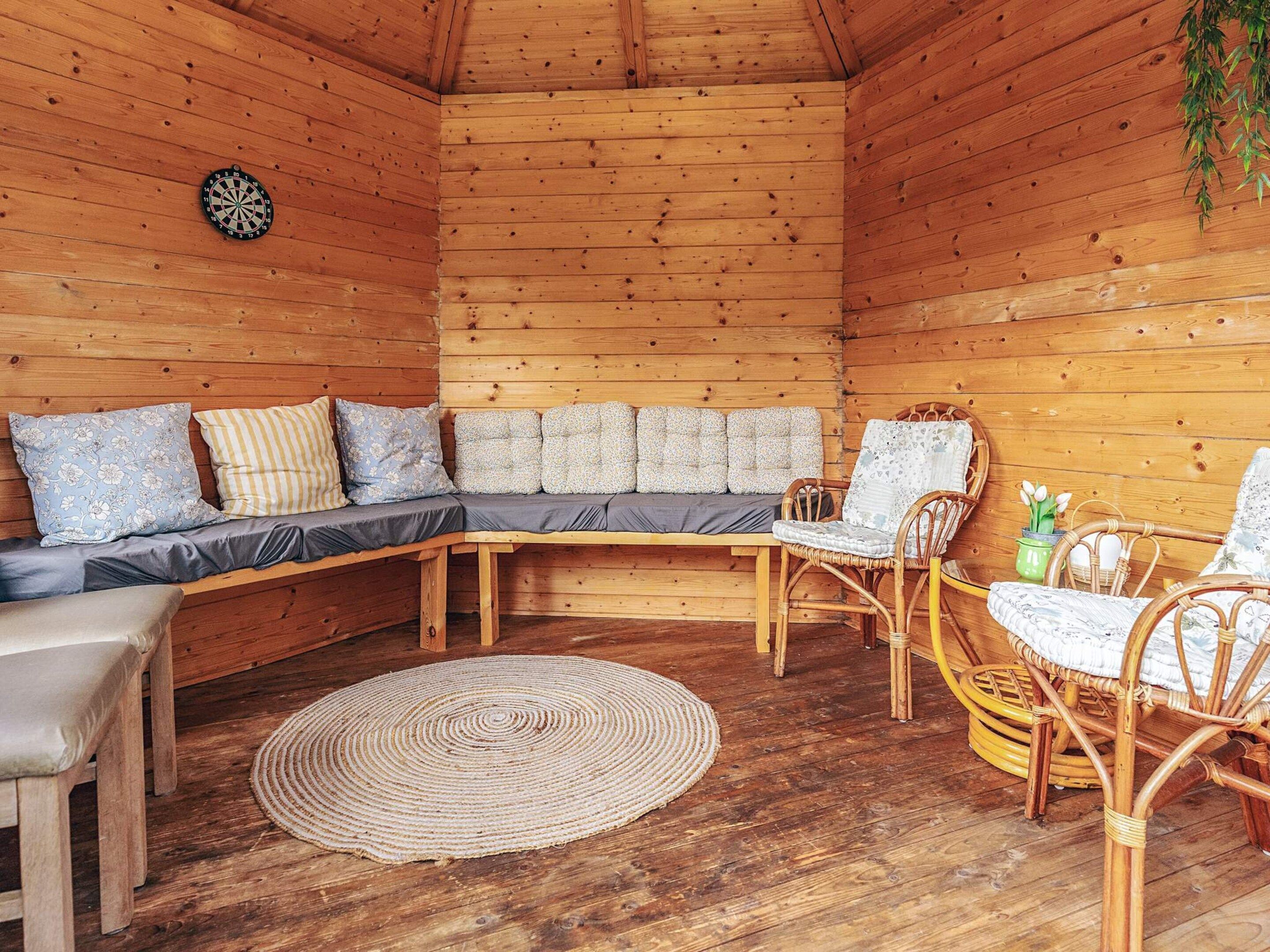 Interior acolhedor de uma cabana de madeira, com um sofá seccional, cadeiras de vime, um tapete tecido e um alvo de dardos.