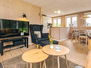 A modern living room with a large flat-screen TV, a black leather armchair, a wooden dining table, and a kitchen area visible through an open door. - Lovely Holiday Home in LÃ¸nstrup near Sea (Hjørring)