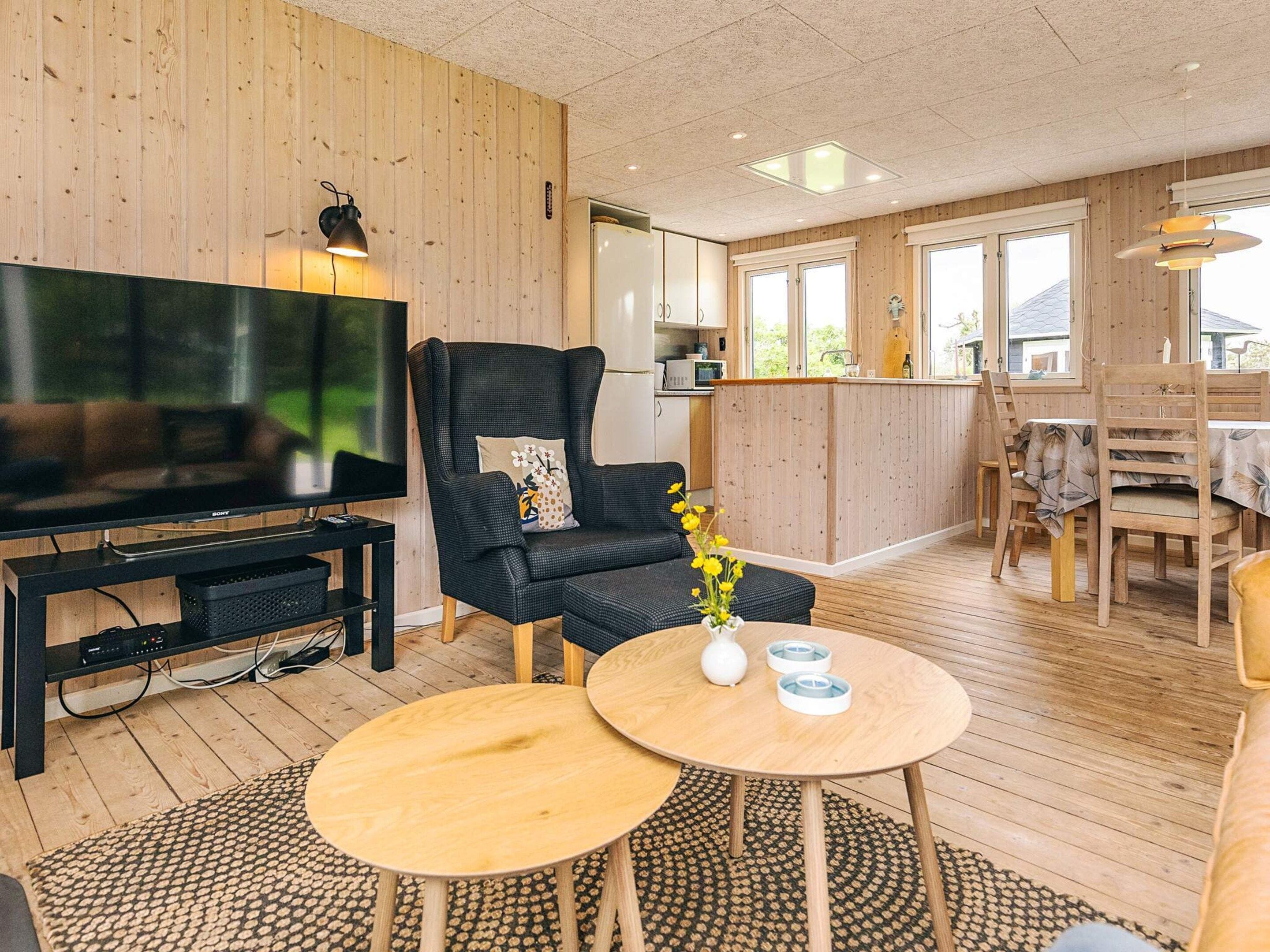 A modern living room with a large flat-screen TV, a black leather armchair, a wooden dining table, and a kitchen area visible through an open door.