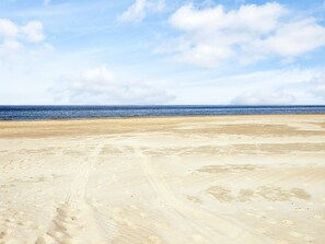 Ein weiter Sandstrand mit Fußabdrücken und Radspuren, der zum Meer unter einem teils bewölkten Himmel führt.