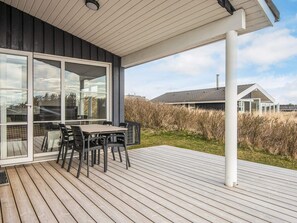 A wooden deck with a table and chairs, large sliding glass doors, and a view of a house and vegetation.