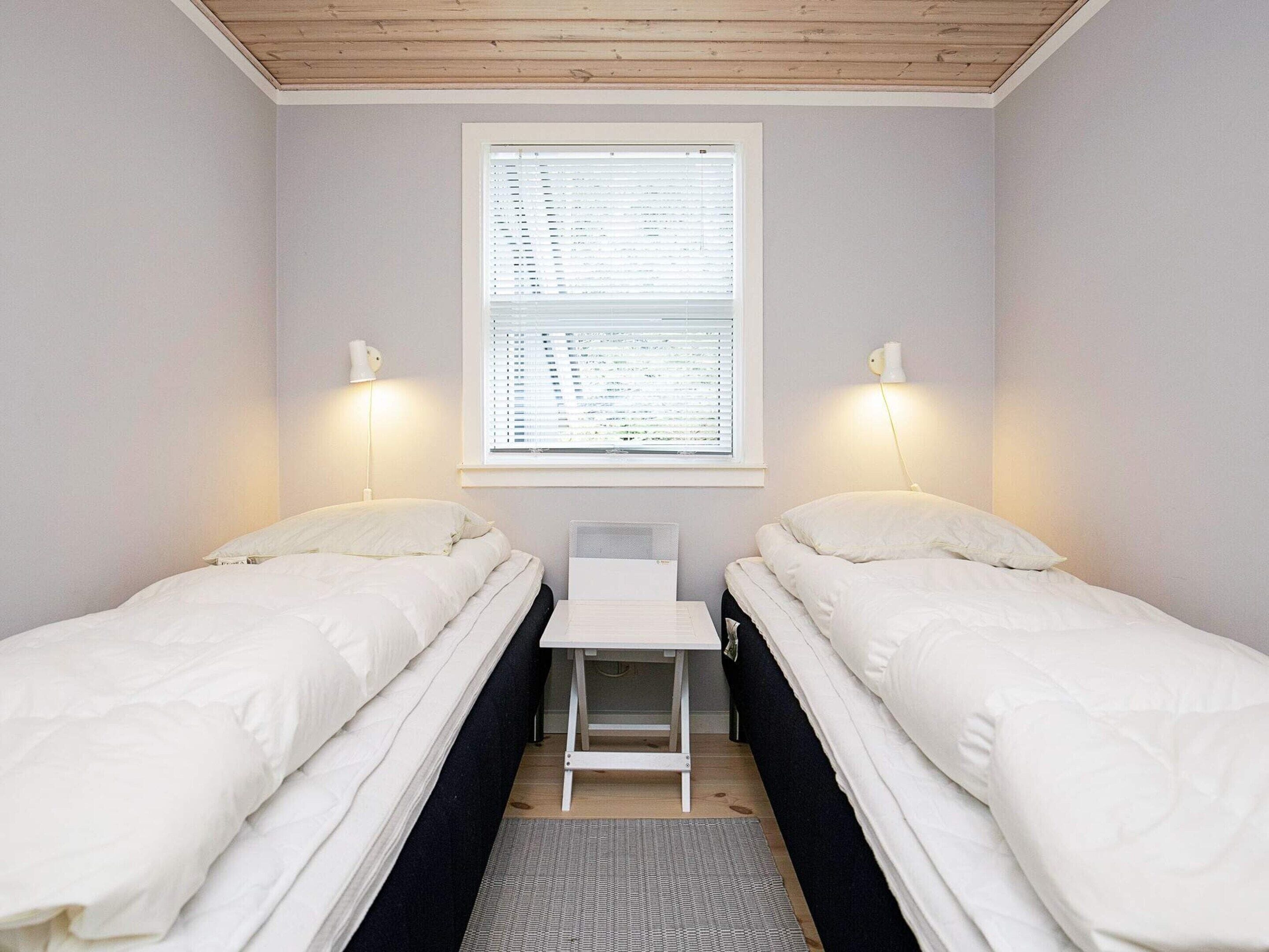 A small, single-room accommodation with two beds, a window with blinds, and a desk.