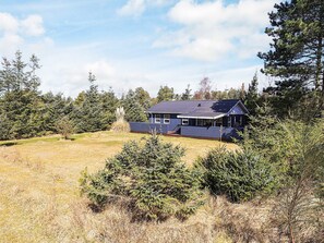Une maison de plain-pied à l’extérieur bleu foncé, entourée d’un champ herbeux et d’une forêt dense.