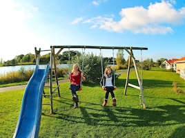 A playground with swings and a slide, set against a backdrop of a lake and houses.