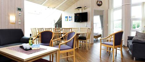 A spacious living area with a wooden ceiling, a dining table with chairs, a sofa, and a television mounted on the wall.