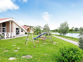 Un terrain de jeux avec un toboggan et des balançoires, une maison avec un toit rouge et un sentier longeant une étendue d’eau.